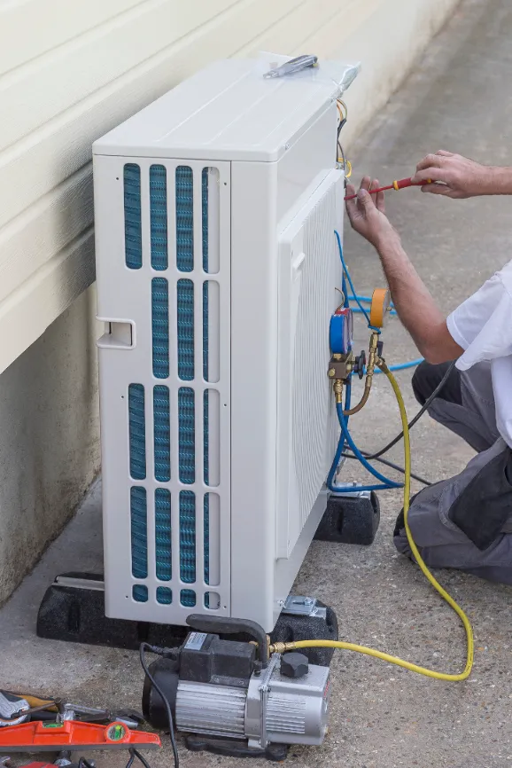 A person uses tools and gauges to service or install an outdoor air conditioning unit next to a building.