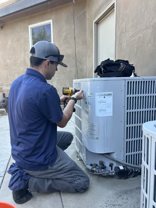 A technician in a blue shirt and hat uses a power drill to service an outdoor air conditioning unit next to a building as part of a routine maintenance plan.