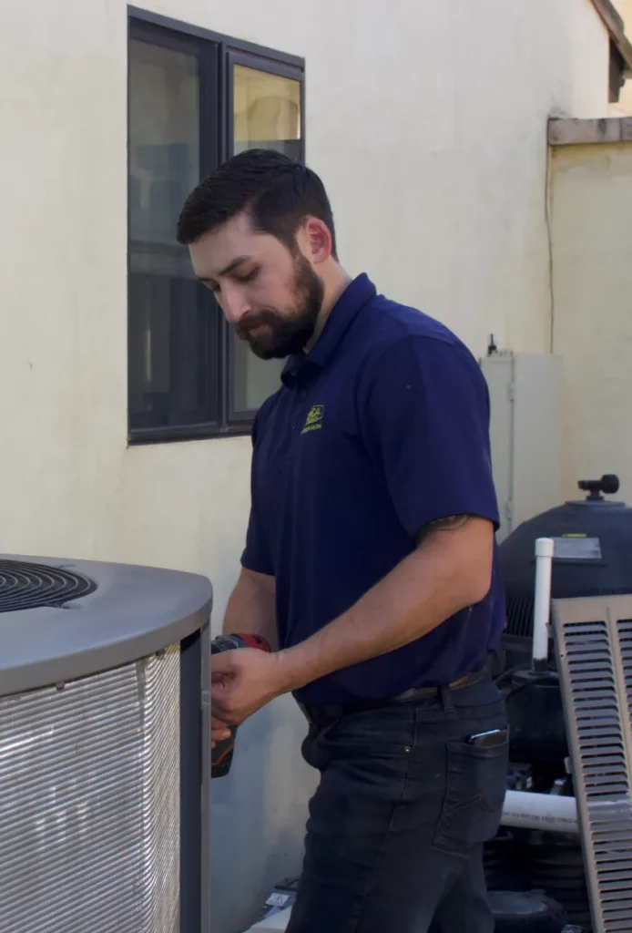 A technician in a blue shirt repairs or maintains an outdoor air conditioning unit next to a building, ensuring the system runs smoothly as part of a regular maintenance plan.