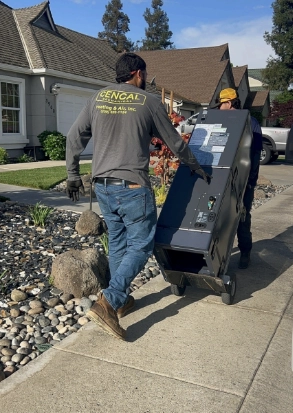 Two workers use a hand truck to move a large HVAC unit along a residential sidewalk in front of a house, making upgrades easy with available financing options.