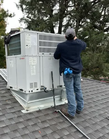 A worker in jeans and a hoodie follows a maintenance plan while servicing a large rooftop HVAC unit surrounded by trees on an overcast day.
